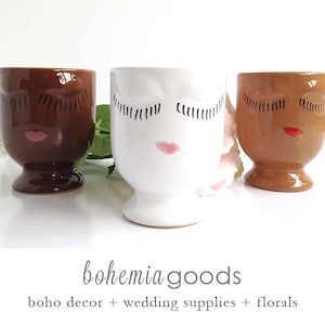 May include: Three ceramic face planters in shades of brown and white. Each planter features a face design with closed eyes and painted lips. The planters are on a white surface. The text "bohemia goods" is at the bottom.