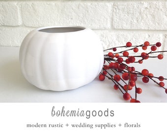 May include: A white, pumpkin-shaped ceramic vase with a gray interior. Red berry branches are placed to the right of the vase. The text "bohemiagoods" is below the vase, with "modern rustic + wedding supplies + florals" underneath.