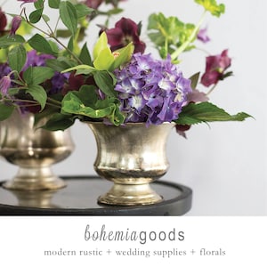May include: Two silver-toned urns filled with vibrant purple hydrangeas, green foliage, and other flowers. The urns sit on a dark surface. The image includes the text "bohemiagoods" and "modern rustic + wedding supplies + florals".