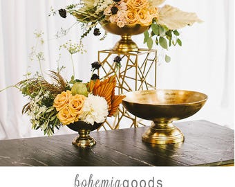 May include: A display of floral arrangements in gold-toned metal bowls. One bowl is on a geometric stand. The arrangements feature orange and white roses, and other dried flowers. The text "bohemiagoods" is at the bottom.