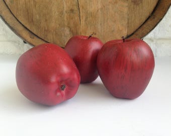 May include: Three red apples with a glossy finish are arranged on a white surface. The apples have a natural appearance, with subtle variations in color and texture. A wooden barrel top is in the background.