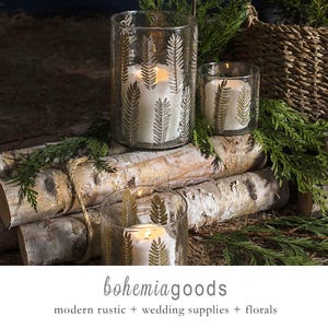 May include: Clear glass candle holders with gold leaf pattern, each holding a white candle. The candle holders are arranged on birch logs with greenery. The text "bohemiagoods" is at the bottom of the image.