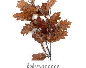 May include: A decorative artificial branch featuring multiple brown oak leaves with a light dusting of white glitter. The text bohemiagoods modern rustic + wedding supplies + florals is printed in grey at the bottom of the image.