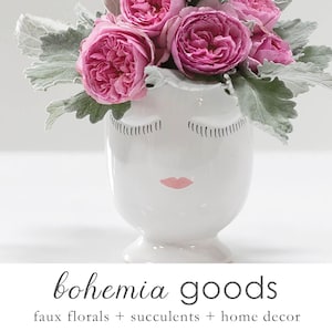 May include: A white ceramic vase with a face design, holding pink roses and gray-green foliage. The vase has closed eyes and a pink lip design. The text "bohemia goods" is at the bottom, with "faux florals + succulents + home decor" below it.