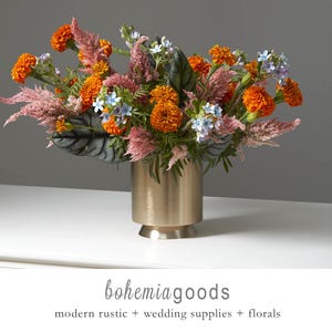 May include: A floral arrangement in a gold-colored vase. The bouquet features orange marigolds, pink feathery plumes, and small blue and white flowers. The vase has a cylindrical shape with a flared base. The text "bohemiagoods" is at the bottom.
