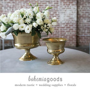 May include: Two gold-colored urns, one filled with white flowers, sit on a round table. The urns have a pedestal base and a wide, shallow bowl. The background features a brick wall. The text "bohemiagoods" is at the bottom.