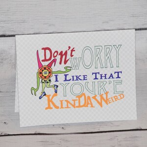 May include: A greeting card with the text "Don't worry I like that you're kinda weird." The card features a cartoon monster with a large eye and a colorful design. The background has a subtle diamond pattern.