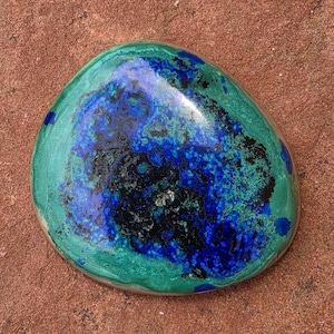 May include: A polished, rounded stone featuring a vibrant mix of deep royal blue, bright turquoise green, and dark black mineral patterns. The surface has a smooth, glossy finish that highlights the intricate, swirling color variations throughout.