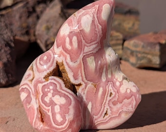 May include: A polished, freeform rhodochrosite stone with a swirling pattern of pink and white bands. The stone has a natural, organic shape and a smooth, glossy surface. The rhodochrosite is a vibrant pink color with white and brown inclusions.