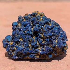 Op de afbeelding: Een ruw mineraal exemplaar met een cluster van diepblauwe, kristallijne formaties met een getextureerd, hobbelig oppervlak. De donkerblauwe kristallen zijn doorspekt met vlekken van aardse, mosterdgele mineraalafzettingen in de structuur.