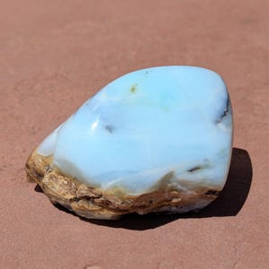 May include: A polished, natural stone with a smooth, light blue surface. The stone has a rounded shape with a brown, textured base. The blue color has subtle variations and a glossy finish, suggesting it is a gemstone or mineral specimen.