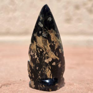 May include: A polished stone carving shaped like a pointed arrowhead or flame. The object features a deep black base color with prominent, irregular patches of metallic gold and bronze inclusions scattered across its smooth, reflective surface.