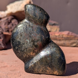 May include: A dark, abstract sculpture with a smooth, curved form. The stone has a metallic sheen with shades of black, gold, and red. The sculpture is displayed on a reddish-brown surface, with blurred background elements.