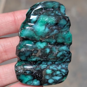 May include: A polished, irregularly shaped gemstone featuring a vibrant teal and green color palette with prominent, dark, web-like matrix patterns throughout. The stone has a smooth, glossy surface with distinct horizontal grooves along the sides.