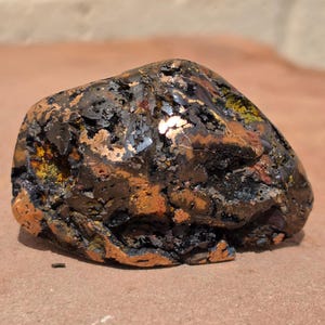 Könnte beinhalten: Ein rohes Mineralexemplar mit einer komplexen, unregelmäßigen Oberfläche und metallischem Glanz. Der Stein zeigt eine Mischung aus tiefem Schwarz, Dunkelbraun und leuchtenden kupferfarbenen Flecken mit sichtbaren porösen Texturen und Kristallbildungen.