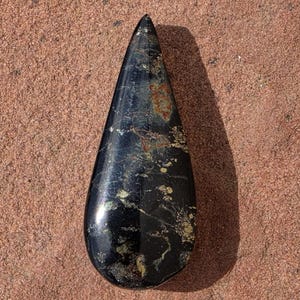 May include: A polished, teardrop-shaped stone cabochon featuring a deep black base colour with scattered metallic gold and bronze-toned mineral inclusions throughout its smooth, reflective surface, tapering to a point at the top end.
