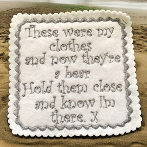May include: A white felt coaster with a scalloped border and embroidered text in silver thread. The text reads "These were my clothes and now they're a bear. Hold them close and know I'm there. X".