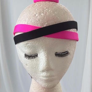 May include: A mannequin head with black and cerise headbands. The headbands are crisscrossed. The mannequin has eyelashes with rhinestones and a cerise top. The background is white.