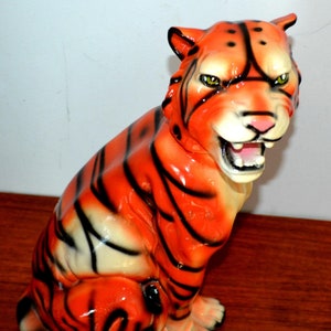 Può includere: Una statuetta di tigre in ceramica con strisce arancioni e nere. La tigre è seduta con la bocca aperta, mostrando i denti. La statuetta è dipinta con una finitura lucida.