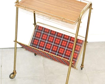 May include: A vintage serving cart with a wooden top and a red and black plaid magazine rack. The cart has a gold-colored metal frame and rolls on small wheels. A stylish piece for entertaining or displaying items.