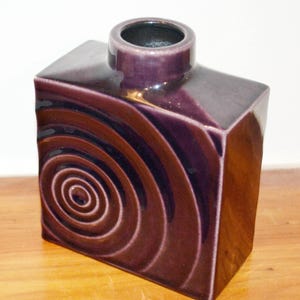 Könnte beinhalten: Eine quadratische Keramikvase in Lila- und Brauntönen. Die Vase hat ein Spiralmuster auf einer Seite und eine zylindrische Öffnung an der Oberseite. Die glänzende Oberfläche reflektiert das Licht und hebt die geometrische Form und das dekorative Muster hervor.