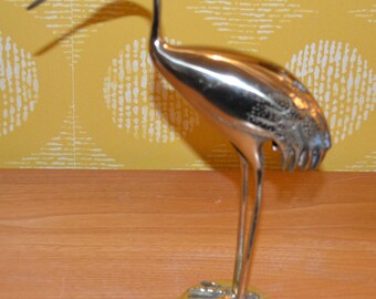 Op de afbeelding: Een zilveren metalen kraanvogelbeeldje met een lange nek en gedetailleerde veren. De kraanvogel staat op een ronde basis met een getextureerd oppervlak.