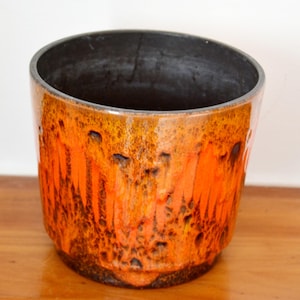 Puede incluir: Una maceta de cerámica con un esmalte brillante naranja y marrón. El interior es oscuro y el borde es de color gris oscuro. La maceta tiene una superficie texturizada y es adecuada para uso en interiores o exteriores.