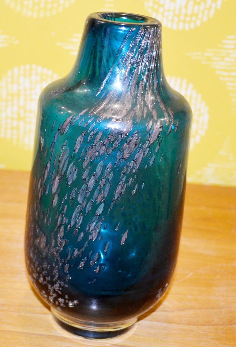 May include: A teal blue glass vase with silver flecks throughout. The vase has a round body and a narrow neck.