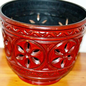 May include: A red decorative planter with a carved floral design. The planter has a dark interior and a wide rim. The exterior features repeating circular patterns with flower-shaped cutouts. The planter is made of a glossy material.