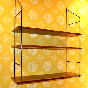 May include: A three-tiered wall-mounted shelf unit. The shelves are made of wood and are a warm brown color. The supporting structure is black metal. The shelves are mounted on a yellow background with a white circular pattern.