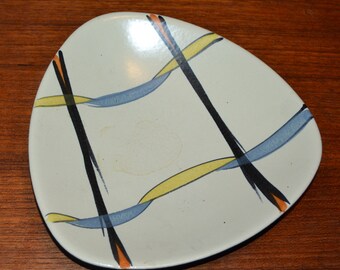 Op de afbeelding: Een wit keramiek bord met een driehoekige vorm en een abstract ontwerp met zwarte, gele, blauwe en oranje lijnen.