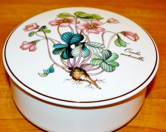 Może przedstawiać: Białe porcelanowe pudełko z pokrywką i wzorem kwiatowym. Pokrywka przedstawia szczegółową ilustrację rośliny z różowymi kwiatami i zielonymi liśćmi. Tekst "Oxalis acetosella" jest nadrukowany na pokrywce czarnym kolorem.