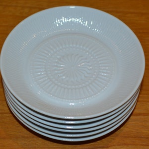 Könnte beinhalten: Ein Stapel von sechs weißen Untertassen aus Keramik mit einem erhabenen, geprägten Design in der Mitte. Die Untertassen sind ordentlich gestapelt, wobei die oberste Untertasse die meisten Details des Designs zeigt.