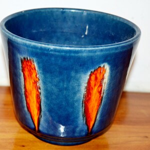 Pode incluir: Um vaso de cerâmica azul com dois desenhos verticais em forma de chama laranja e branca. O vaso tem uma borda ligeiramente angulada e um acabamento brilhante. O design contrasta com o fundo azul.
