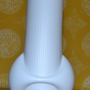 May include: A white ceramic vase with a unique, abstract design. The vase has a tall, cylindrical body with a rounded base and a textured, ribbed surface.