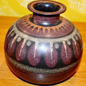 以下が含まれることがあります： 茶色の陶器製の壺で、幾何学模様が施されています。壺は口が広く、丸みを帯びた胴体を持っています。模様は、茶色、白、黒の濃淡で、同心円と三角形が幾重にも重なっています。