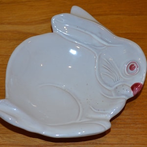 May include: A white ceramic rabbit-shaped dish with a pink nose and eye. The dish is perfect for holding small items like jewelry or keys.