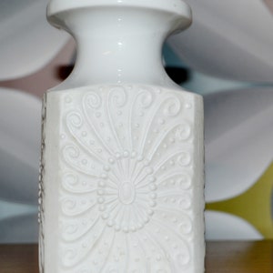 Könnte beinhalten: Weiße Keramikvase mit quadratischem Korpus und einem erhabenen Blumendesign. Die Vase hat einen schmalen Hals und eine breite Öffnung.