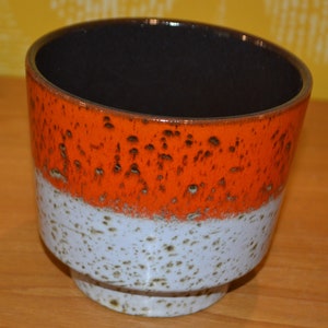 Könnte beinhalten: Ein Keramiktopf mit schwarzem Rand, orangefarbener Glasur und weißer Glasur mit braunen Sprenkeln.