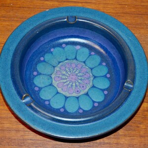 以下が含まれることがあります： 青色のセラミック製の灰皿で、青、紫、白の色の花柄が描かれています。灰皿は幅広の縁と深い穴があります。