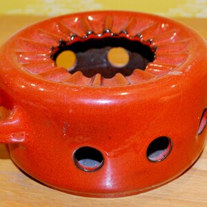 Può includere: Un bruciatore per piano cottura in ceramica rossa con un bordo scanalato e quattro fori. Il bruciatore ha due maniglie sui lati.