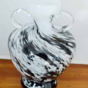 Może przedstawiać: Biały szklany wazon z czarnymi, wirującymi wzorami i dwoma uchwytami. Wazon ma okrągły korpus i rozszerzającą się górę.