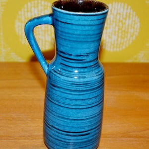 Puede incluir: Un jarrón de cerámica azul con un borde marrón y un asa. El jarrón tiene un patrón de rayas.