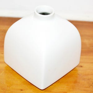 Puede incluir: Un jarrón blanco de forma cuadrada con una parte superior redondeada y una pequeña abertura. El jarrón tiene un acabado liso y brillante y un diseño minimalista. El jarrón está sobre una superficie de madera.