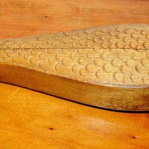 Könnte beinhalten: Ein geschnitztes, fischförmiges Objekt aus Holz mit einer natürlichen hellbraunen Oberfläche. Die Oberfläche weist detaillierte, in das Holz eingearbeitete Schuppenmuster, eine definierte Schwanzflosse und ein kleines kreisförmiges Auge auf.