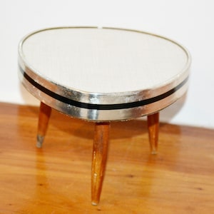 May include: A small, vintage plant stand with a triangular top. The top is white with a silver trim and a black stripe. It has three wooden legs. The stand is approximately 12 inches tall.