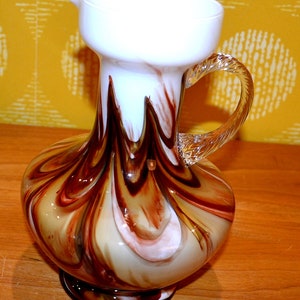 Puede incluir: Una jarra decorativa de vidrio con una parte superior blanca y una base bulbosa. La jarra presenta un patrón arremolinado de colores marrón, beige y blanco. Tiene un asa de vidrio transparente y retorcido y se asienta sobre una base pequeña y redonda.