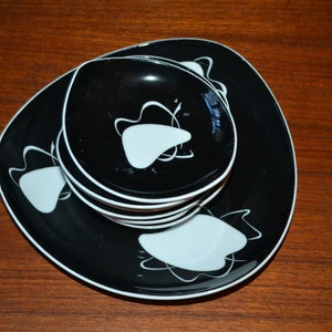 May include: A set of black and white mid-century modern dinnerware. The set includes plates and bowls with a white abstract design and a white rim. The dishes are stacked on a wooden surface.