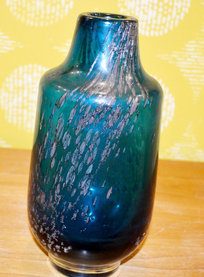 May include: A teal blue glass vase with silver flecks throughout. The vase has a rounded body and a narrow neck.