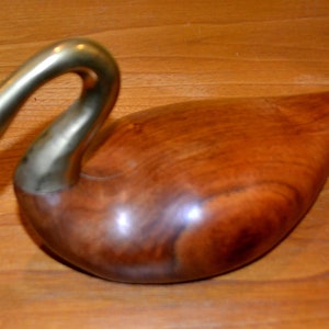 May include: A wooden swan sculpture with a brass neck and head. The swan has a white eye and is resting on a wooden surface.